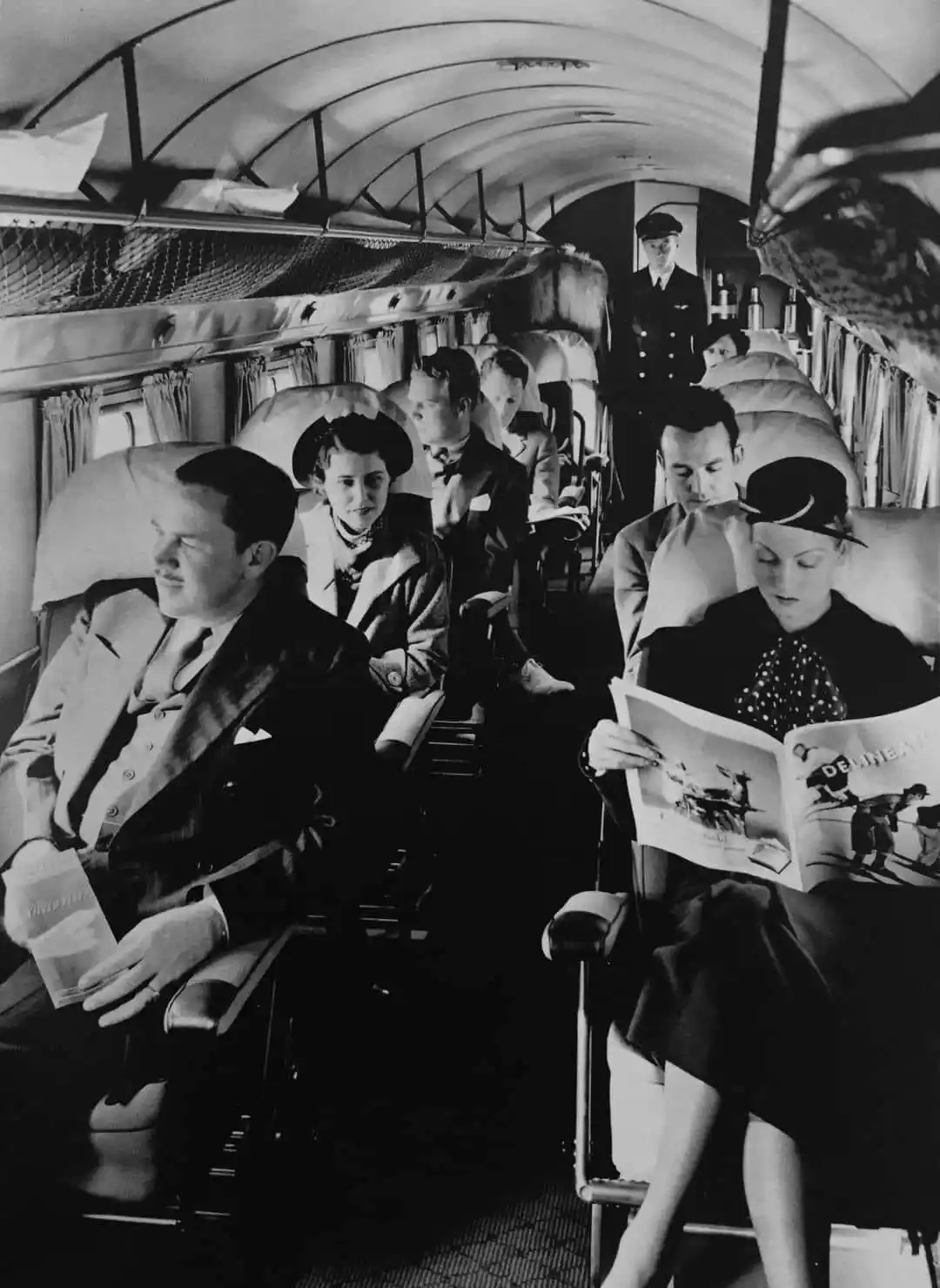 A black-and-white photograph shows passengers seated inside a vintage airplane cabin, dressed in 1940s attire. A woman in…