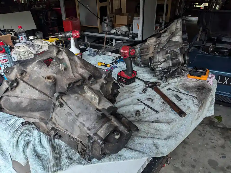 Two dirty, disassembled car transmissions sit on a workbench covered with a towel, surrounded by tools like a red Milwaukee…