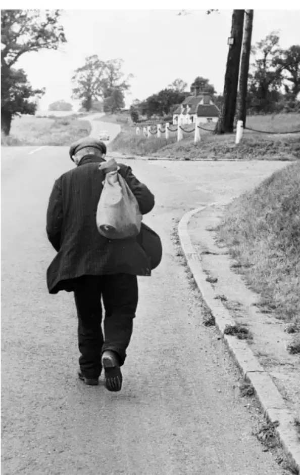 A black-and-white photo shows an older man walking away from the camera down a rural road, carrying a large sack over his…