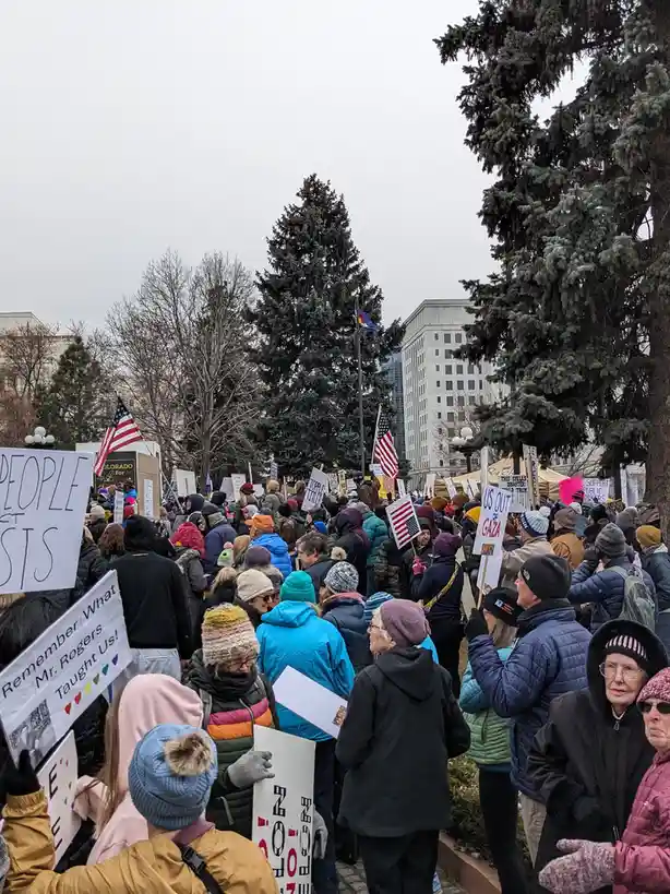 A crowd of people dressed in winter coats and hats holds signs at an outdoor protest, with visible signs reading 'US OUT OF…