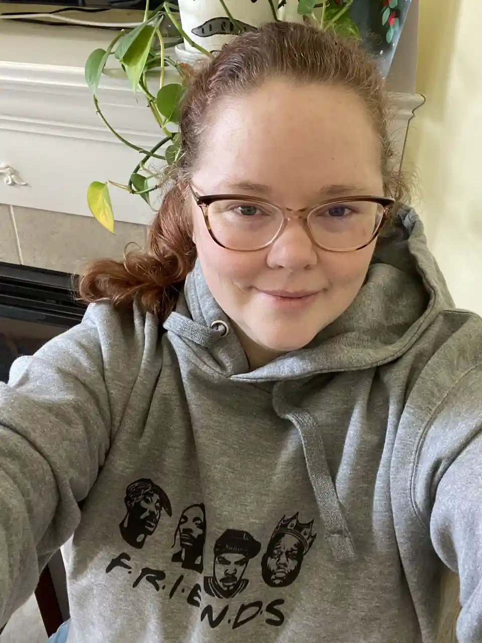 A person with glasses and reddish hair tied back takes a selfie wearing a gray hoodie featuring black silhouettes of four…