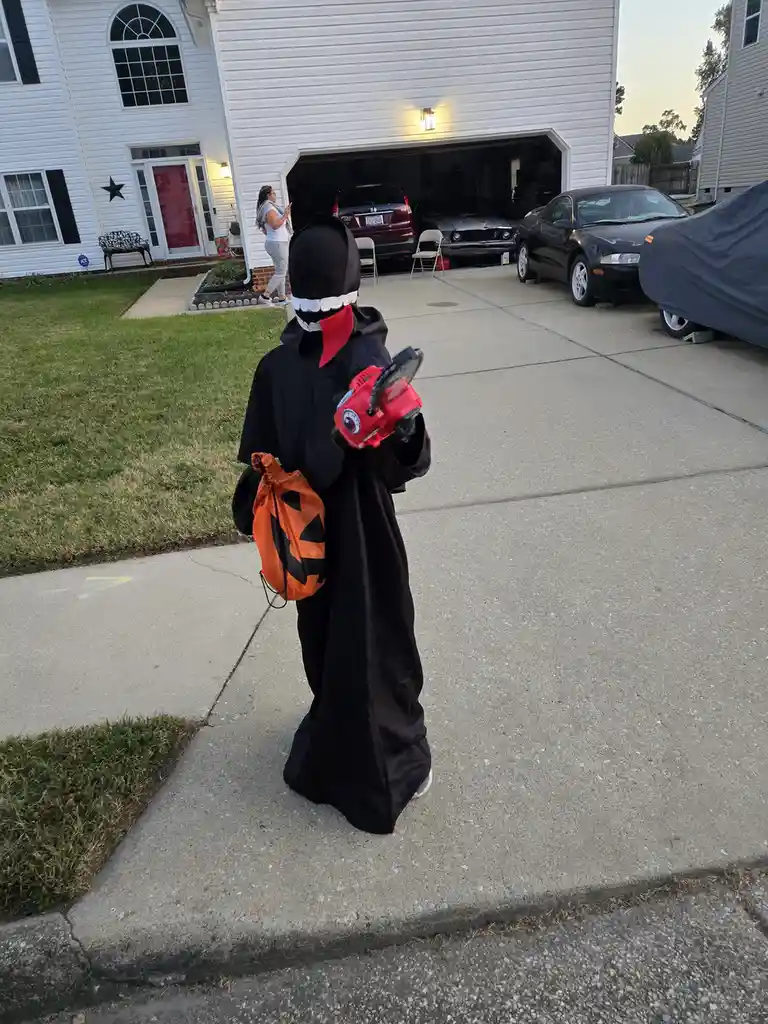 A child dressed in a black Halloween costume with a red scarf and a white-toothed mask walks away from the camera holding a…