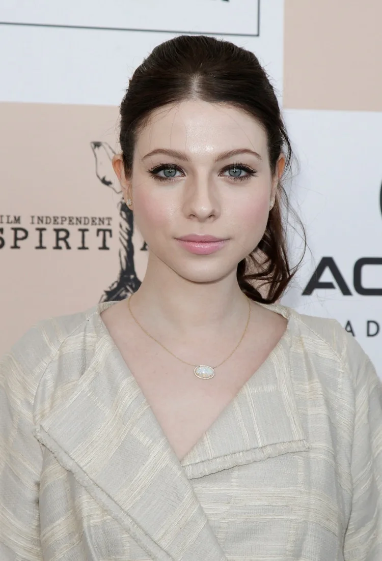 A woman with dark hair pulled back and light makeup looks directly at the camera with a neutral expression. She wears a…