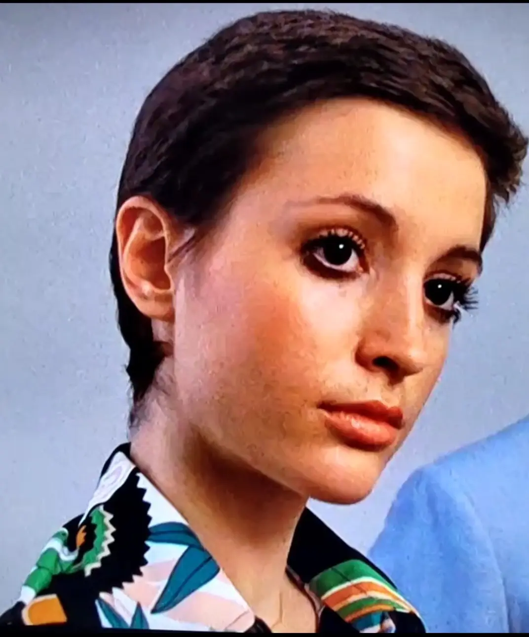 A close-up of a woman with short dark hair and heavy eyelashes, wearing a colorful patterned shirt, looking slightly to the…