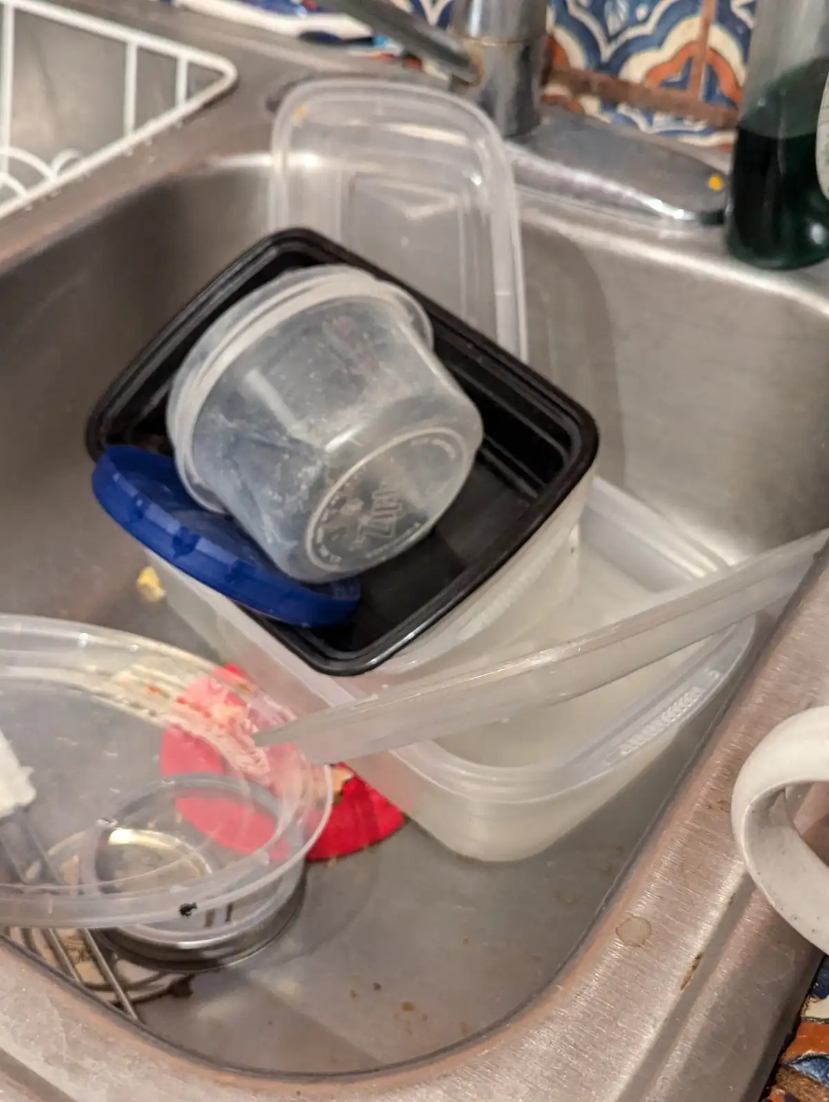 A stainless steel kitchen sink is visibly cluttered with various dirty plastic takeout containers, lids, and food remnants…