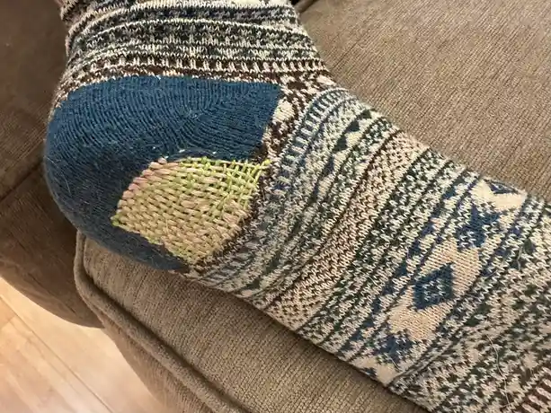 A close-up photo shows a worn, patterned sock with a visible patch sewn onto its heel area, resting on a beige couch. The…