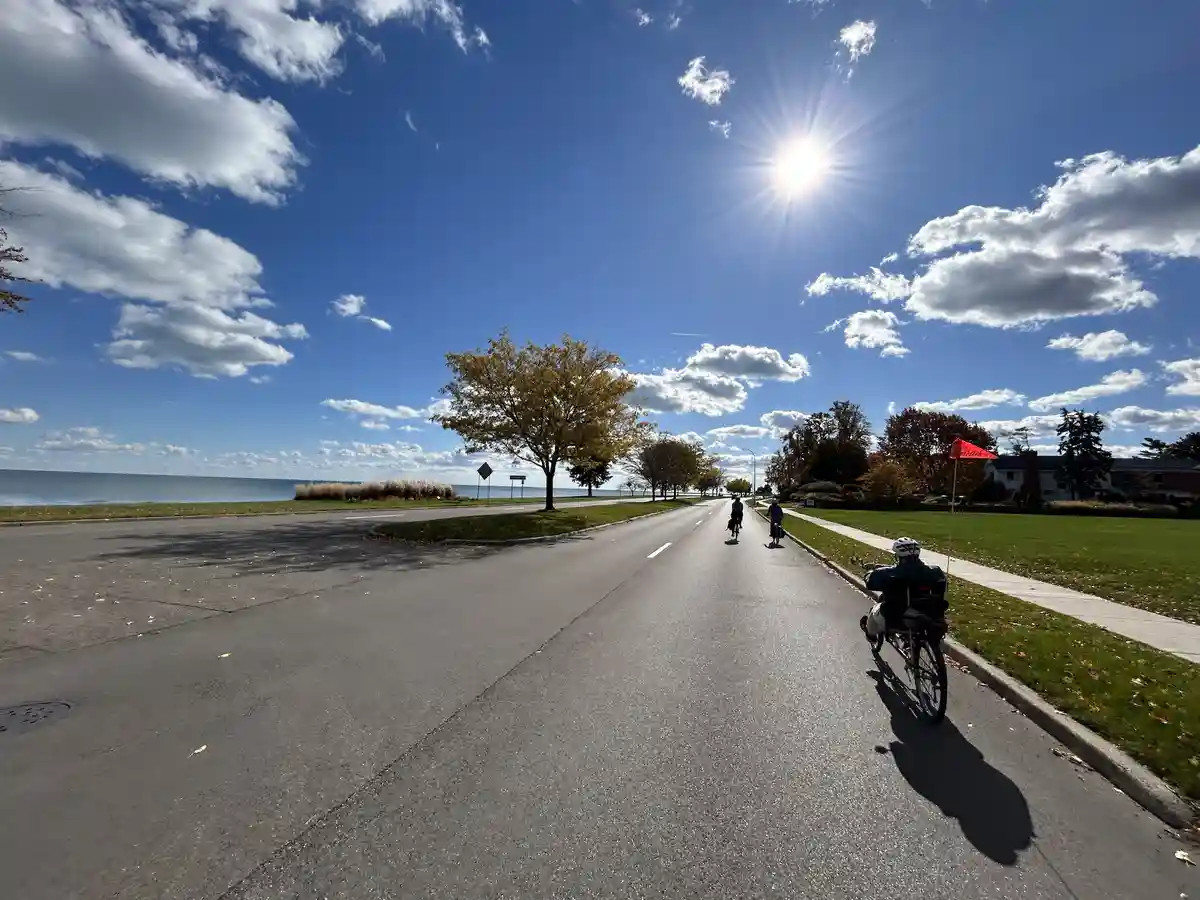 A bright, sunny day with a clear blue sky and scattered clouds. Three cyclists are riding on a paved road next to a grassy…