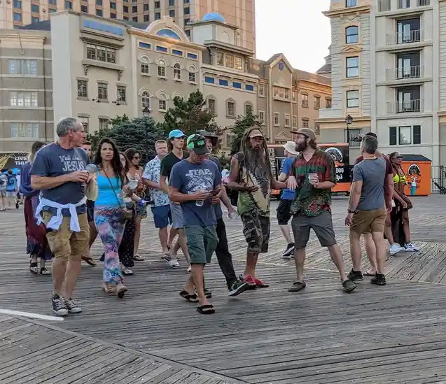 A group of casually dressed people walk along a wooden boardwalk in front of multi-story buildings. One man in the center…