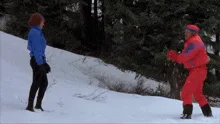 A sequence of six images shows a man in a bright red snowsuit and blue hat attempting to snowboard while another person in a…