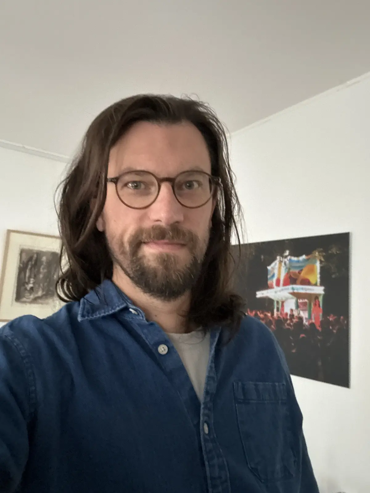 A man with long brown hair, a beard, and round glasses takes a selfie wearing a blue denim shirt over a white t-shirt…