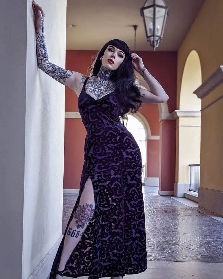A woman with long black hair, heavy makeup, and tattoos on her neck, arms, and thigh poses in a purple gothic-patterned…