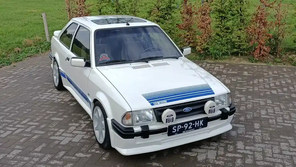 A white Ford RS1600i classic car with blue racing stripes and round headlights is parked on a paved driveway. The license…