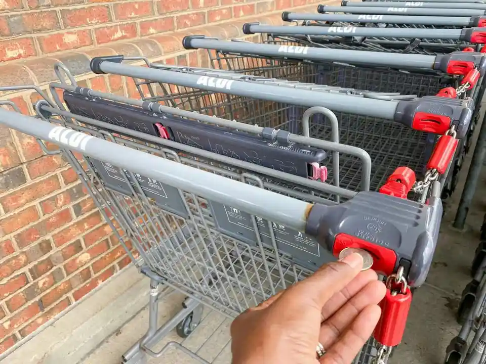 A hand inserts a coin into the lock mechanism of an ALDI shopping cart, which is part of a row of identical gray carts…
