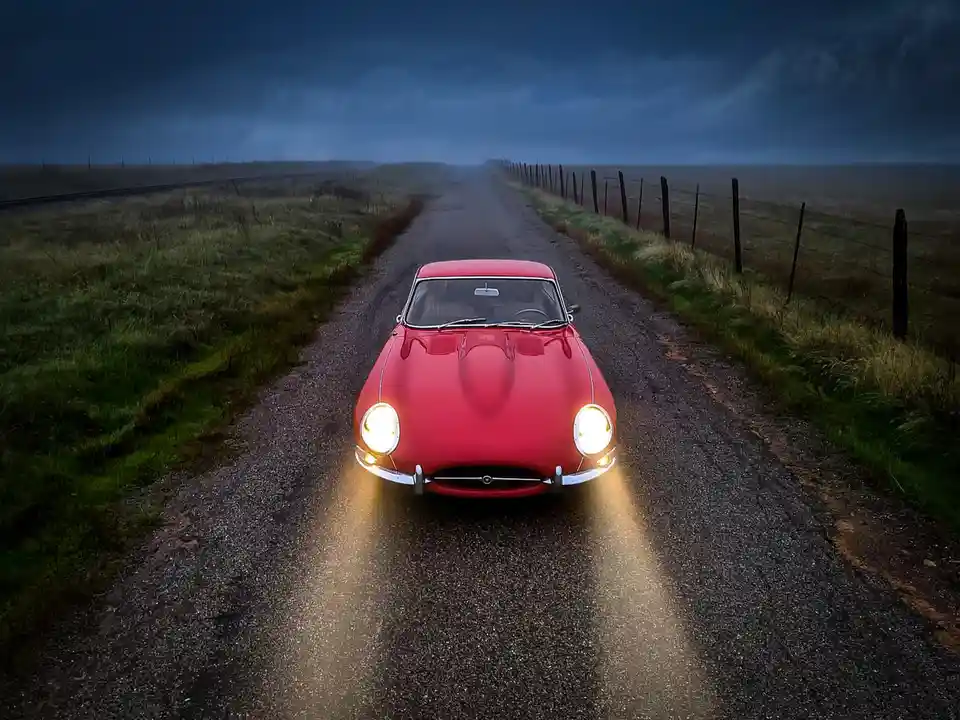 A red vintage sports car with its headlights on drives toward the viewer down a dark, empty rural road under a stormy sky…