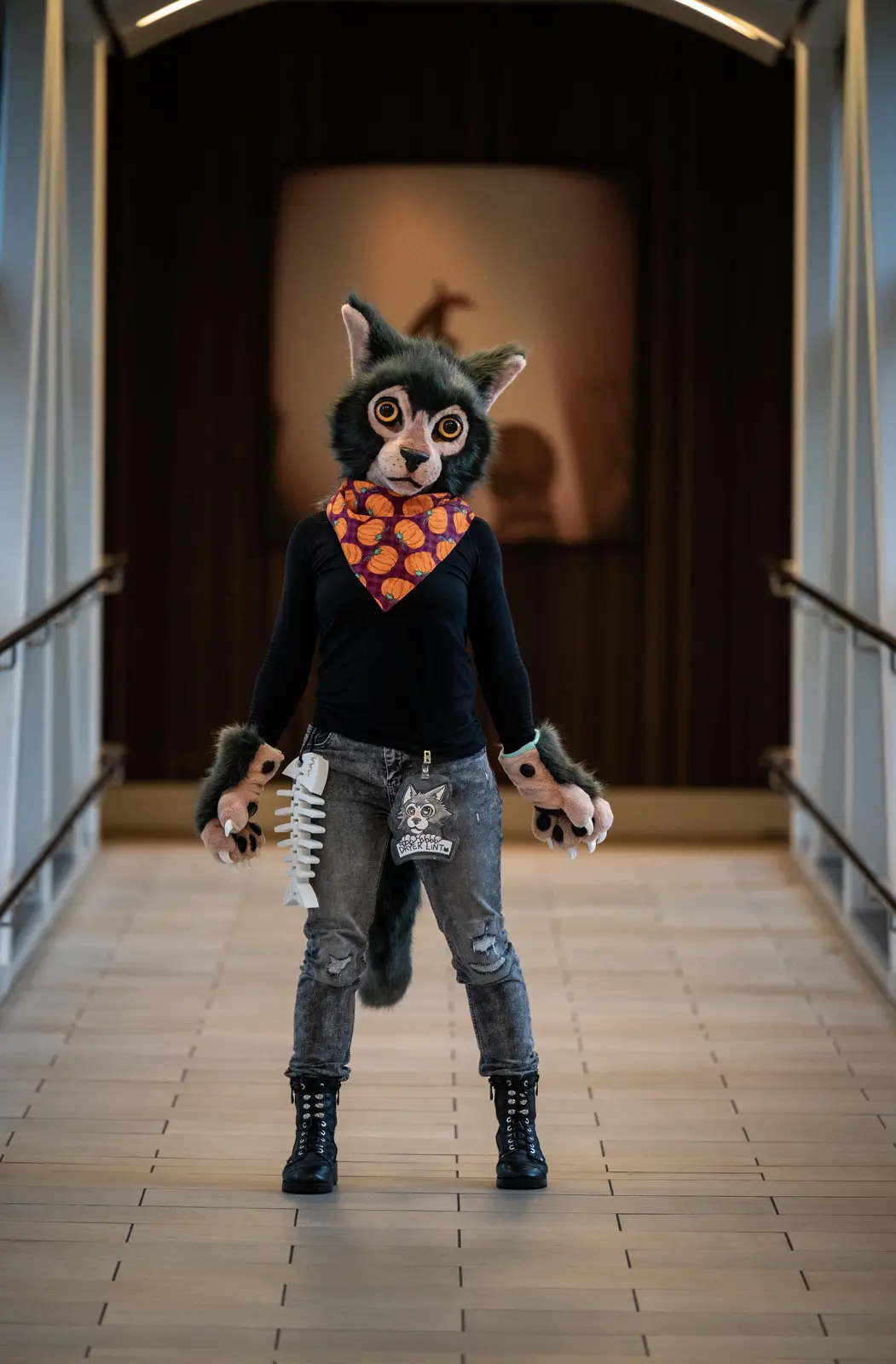 A person in a black-furred wolf-like fursuit stands in a hallway, wearing a black shirt, ripped jeans, and black boots. They…