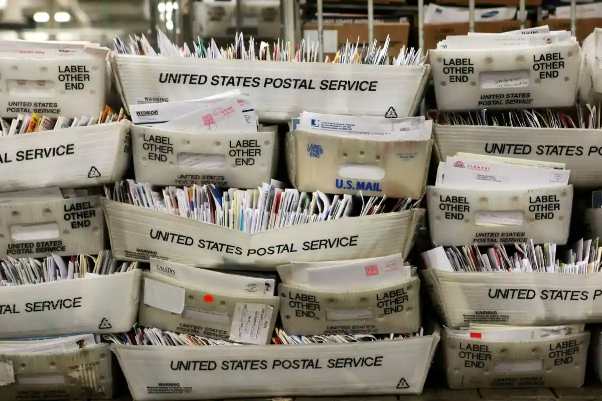 A stack of United States Postal Service cardboard mail trays, overflowing with letters and envelopes, each tray labeled…