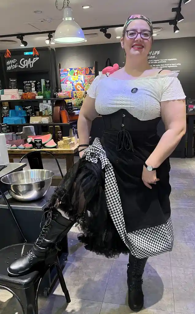 A woman in a store with 'HAND MADE SOAP' signage stands smiling, wearing a white off-the-shoulder top, black corset…