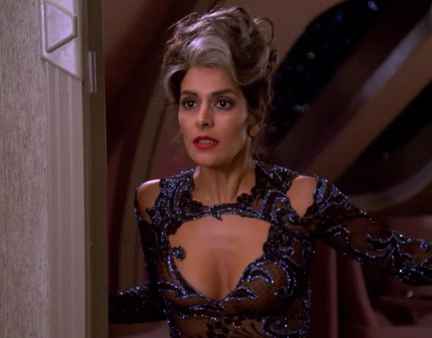 A woman with voluminous, styled hair and red lipstick wears a revealing black beaded dress with cutouts. She stands in a…