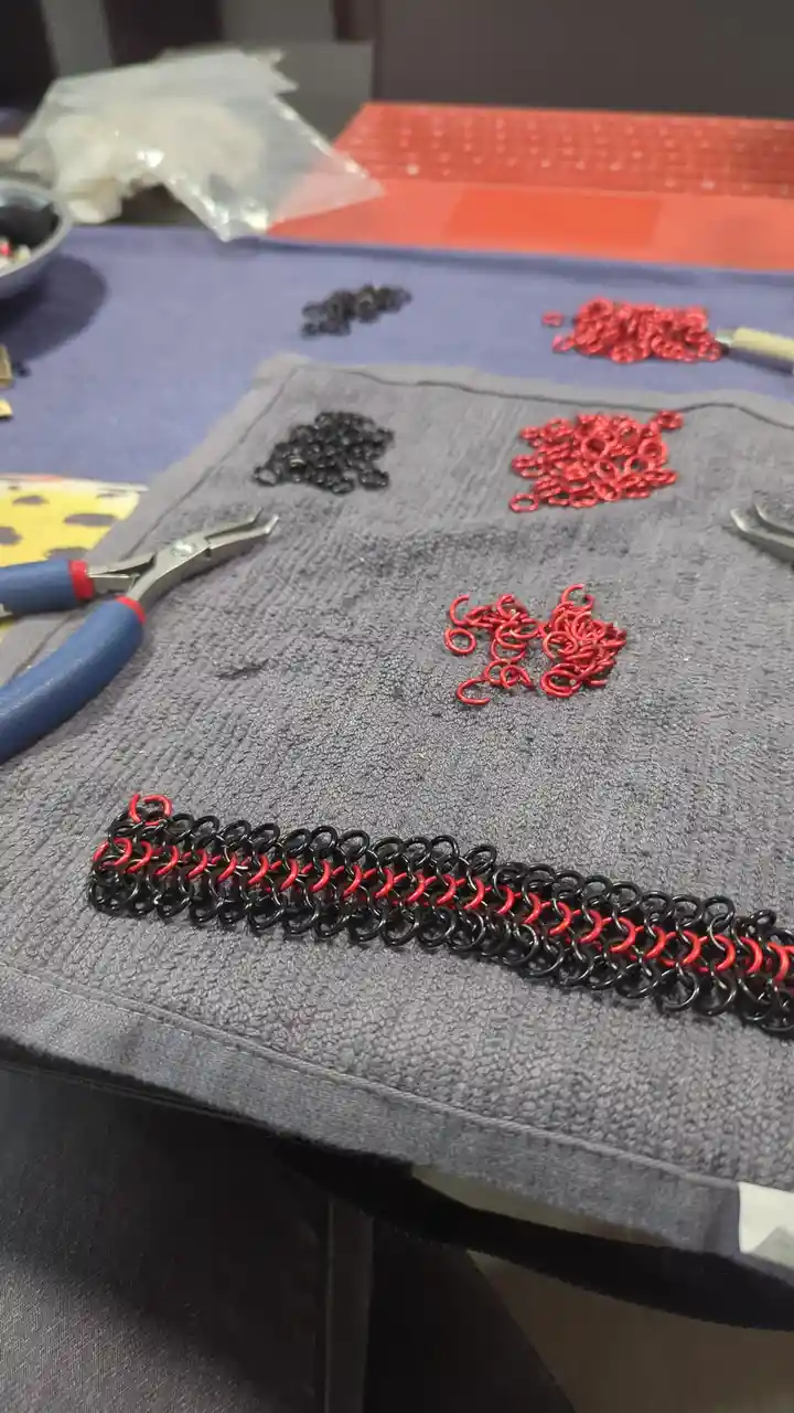 A close-up photo shows a partially completed chainmail bracelet made of interlinked black and red metal rings, resting on a…