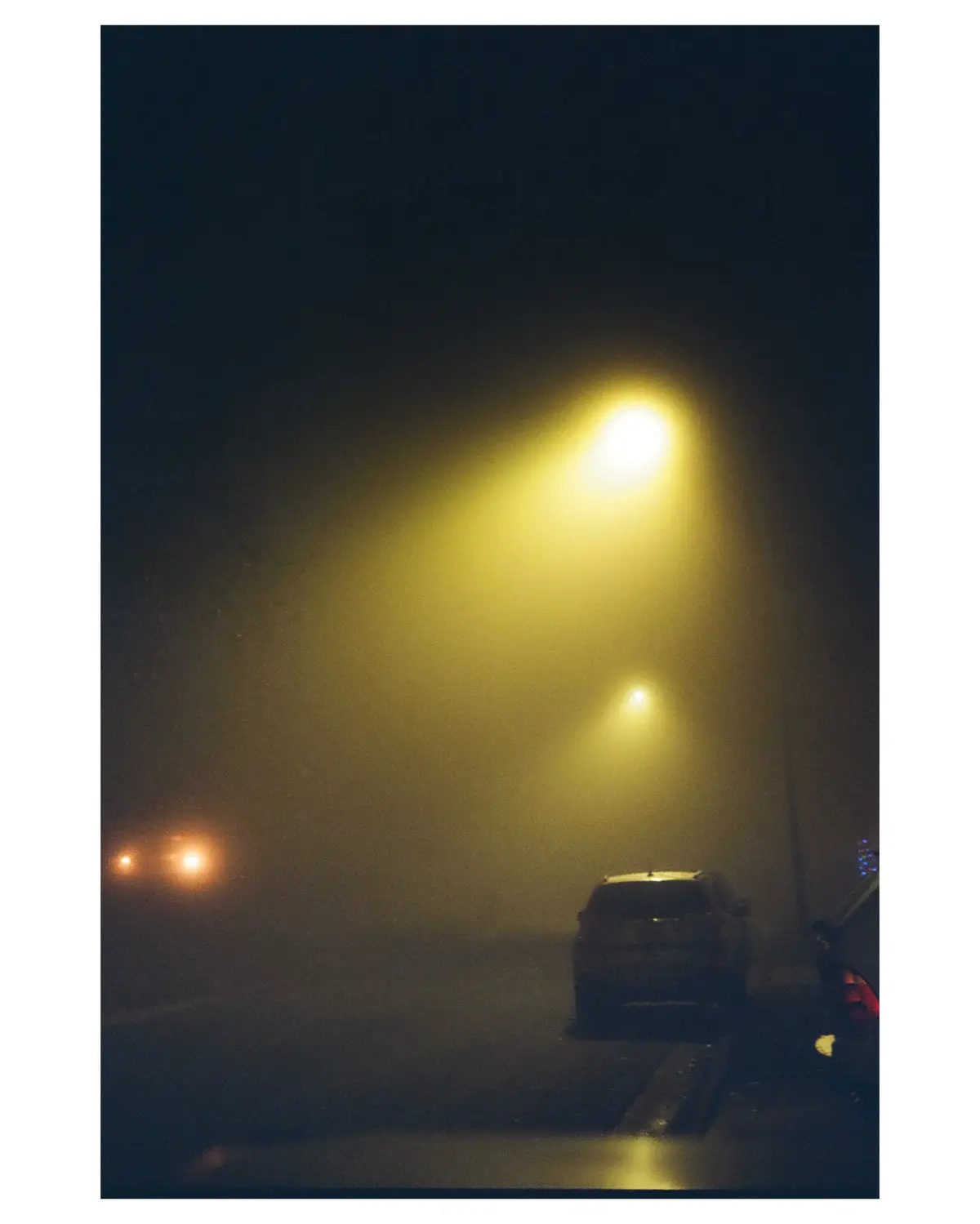 A nighttime photo taken from inside a car shows a foggy street with streetlights casting yellow beams through the haze…