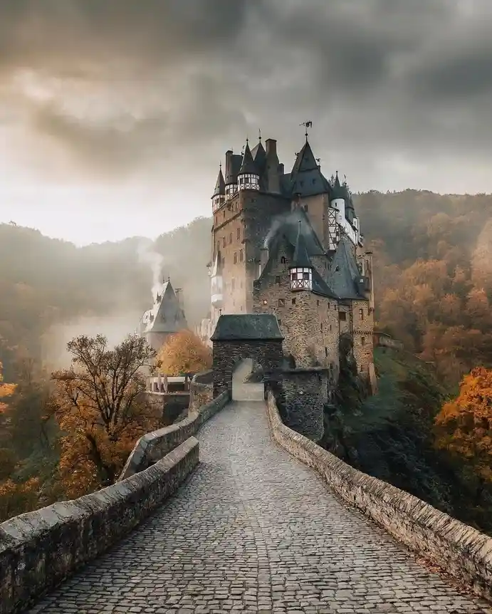A majestic, misty medieval castle with multiple turrets and stone walls sits atop a hill, viewed from a cobblestone bridge…