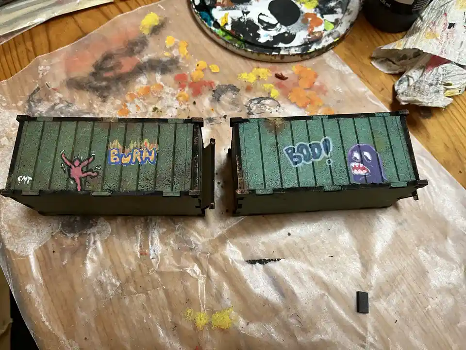 Two small, green-painted model shipping containers sit on a paint-splattered plastic sheet. The left container has a pink…