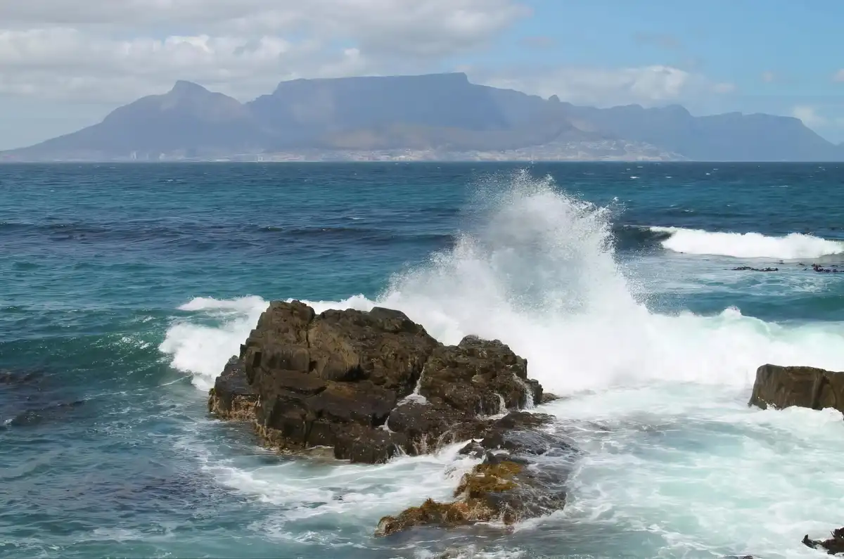 A scenic photo of ocean waves crashing violently against dark rocks in the foreground, with the iconic flat-topped Table…