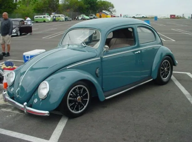 A teal vintage Volkswagen Beetle is parked in a lot with other classic cars in the background. The car has chrome trim…