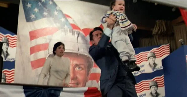 A man in a suit holds a baby high in the air while standing before a large campaign banner featuring a man's face, an…