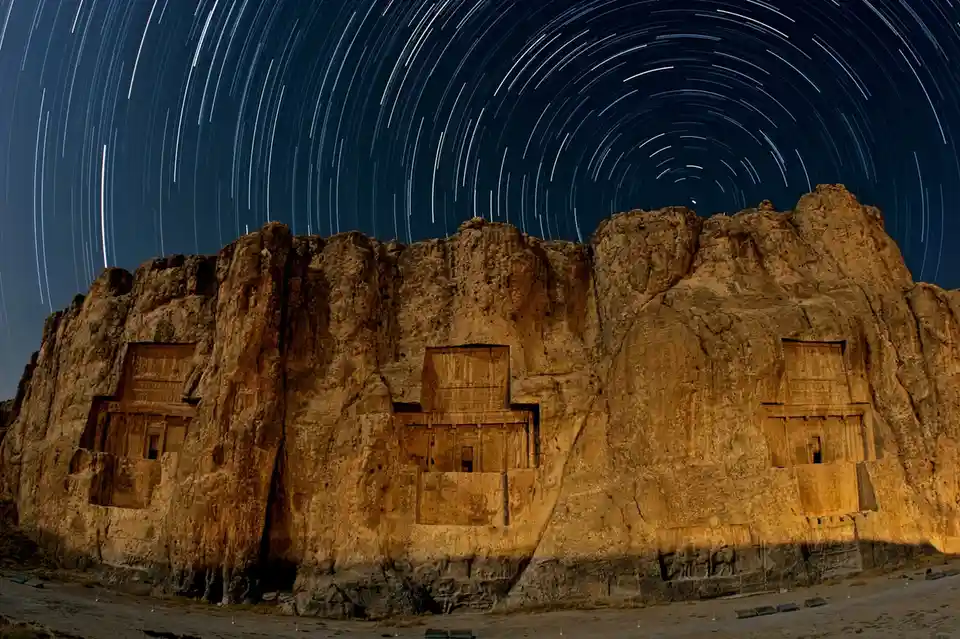 A long-exposure photograph shows ancient rock-cut tombs carved into a cliff face under a night sky filled with bright…