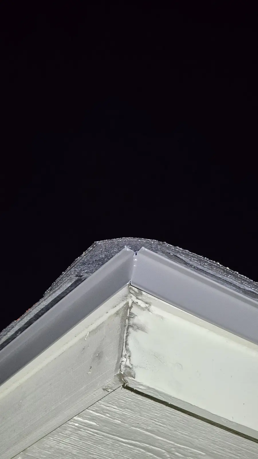 A close-up photo shows the corner of a roof at night, where a gray metal flashing meets a white painted wooden structure…