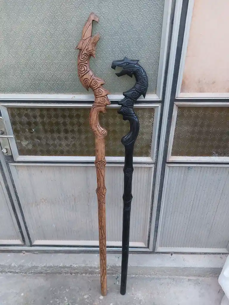 Two intricately carved wooden staffs, one light brown and one black, both featuring dragon-head tops and swirling patterns…
