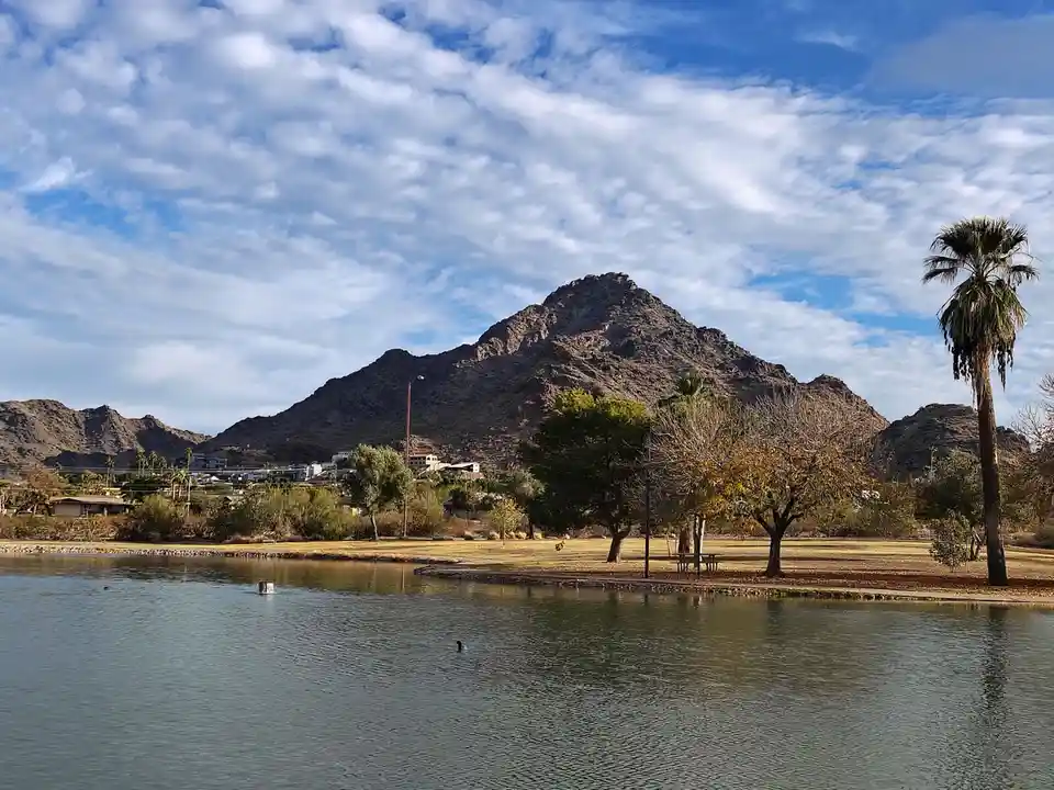 A scenic photo of a calm lake with a large rocky mountain in the background under a partly cloudy sky. A tall palm tree…