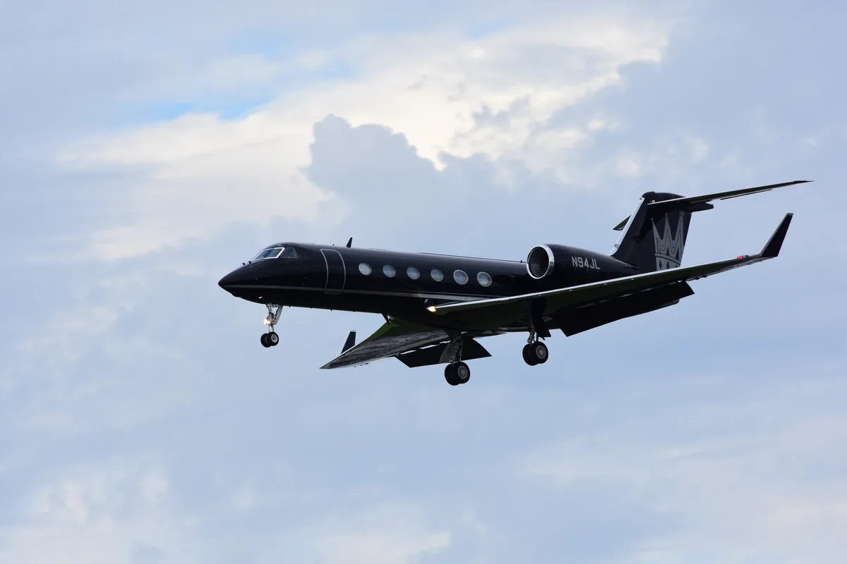A black private jet with landing gear down flies against a cloudy sky, its tail featuring a crown logo and the registration…