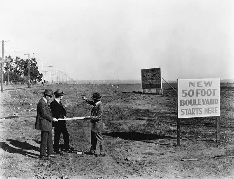 A black-and-white historical photo shows three men in suits and hats standing on undeveloped dirt land. One man points…