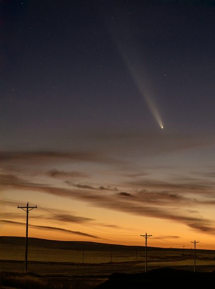 A night sky photo shows a bright comet with a long tail arcing across a starry sky above a rural landscape with power lines…