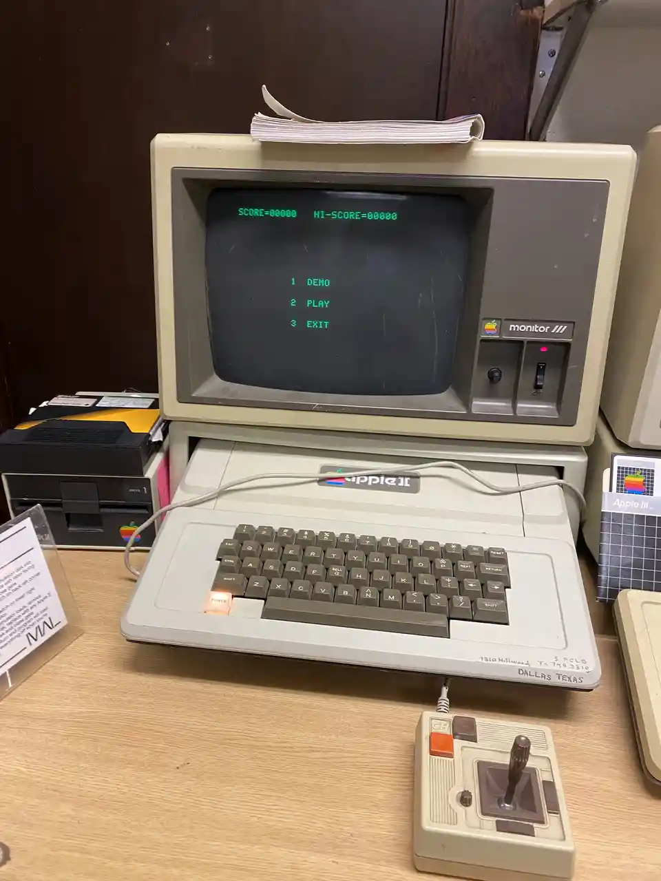 An Apple II computer with a beige monitor and keyboard, displaying a green text menu with options '1 DEMO', '2 PLAY', and '3…
