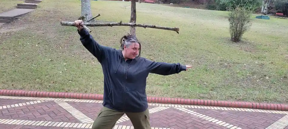 A woman stands outdoors on a patterned brick walkway, holding a long stick horizontally across her shoulders like a weapon…