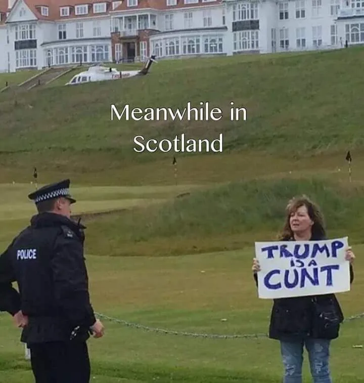 A police officer in a black uniform stands facing a woman holding a handmade sign that reads 'TRUMP IS A CUNT'. The text…