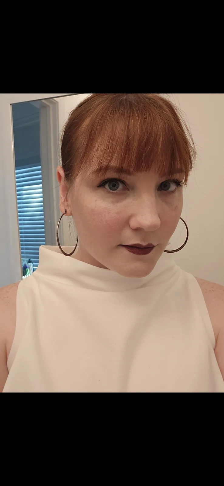 A woman with reddish-brown hair and bangs, wearing large hoop earrings and a white high-neck top, stares directly at the…
