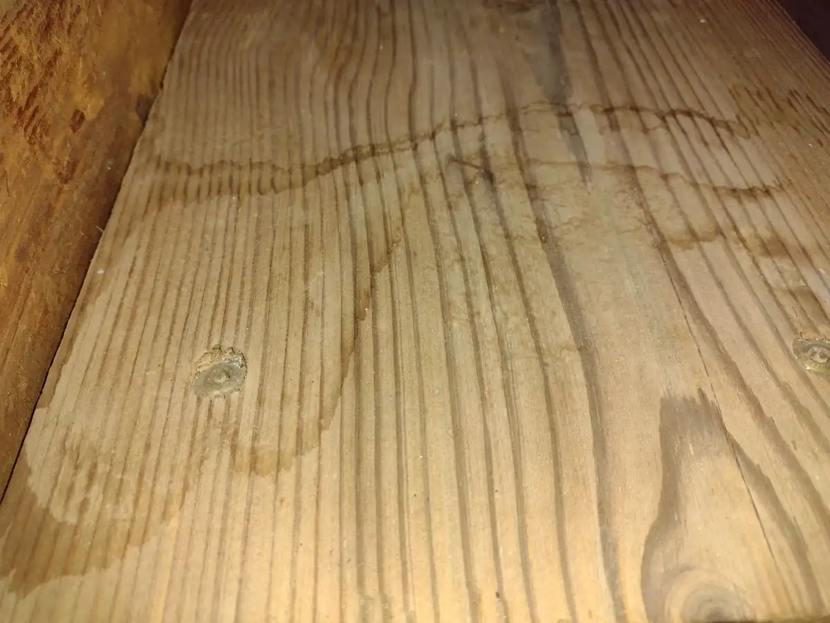A close-up photo of a wooden surface with visible grain, two screw heads, and dark, wavy water stains running across it. The…