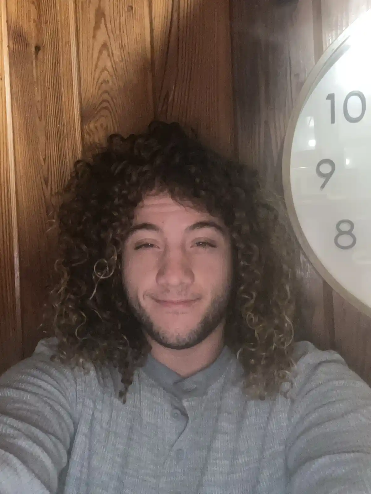 A young man with long curly hair and a light beard smiles smugly at the camera while standing in front of a wooden wall with…