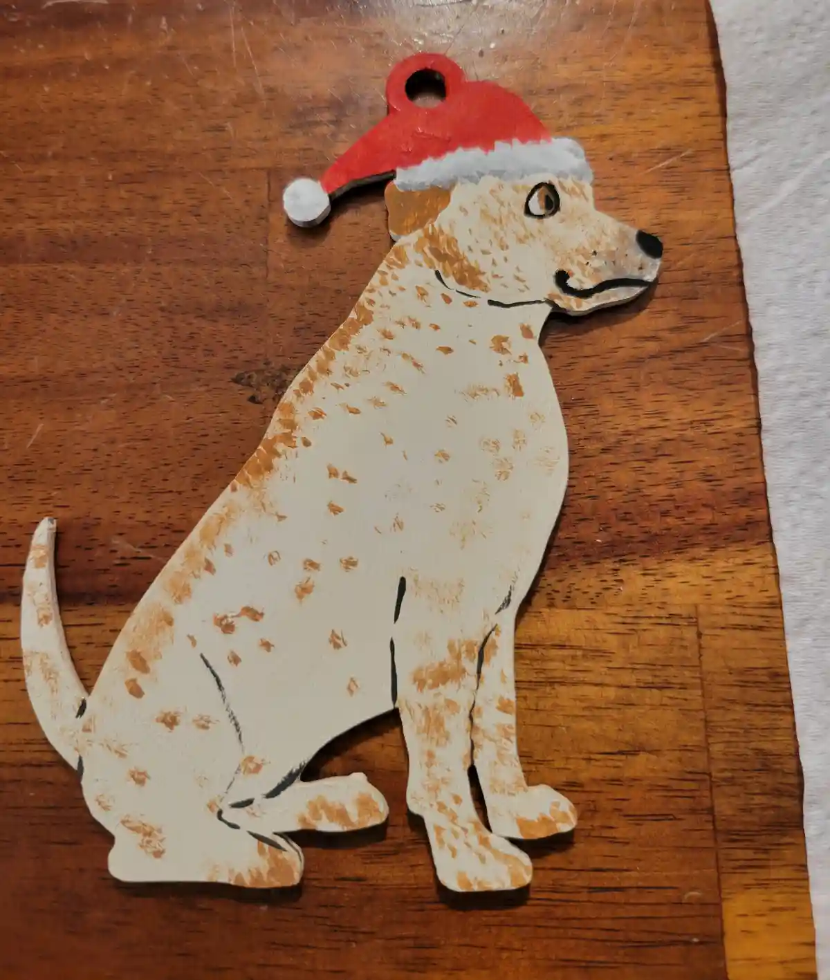 A hand-painted wooden cutout of a seated dog with white and tan speckled fur, wearing a red Santa hat with white trim, rests…