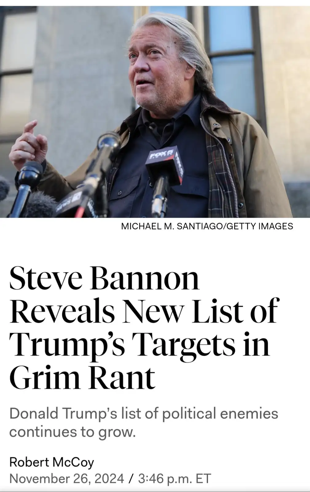 A man with long gray hair, wearing a brown jacket over a dark shirt, speaks into multiple microphones, including one labeled…
