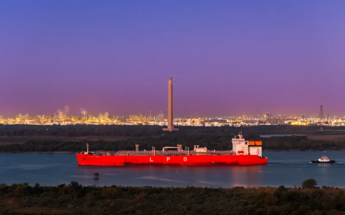 A large red LPG tanker ship is shown on a body of water at dusk, with a city skyline and industrial complex lit up in the…