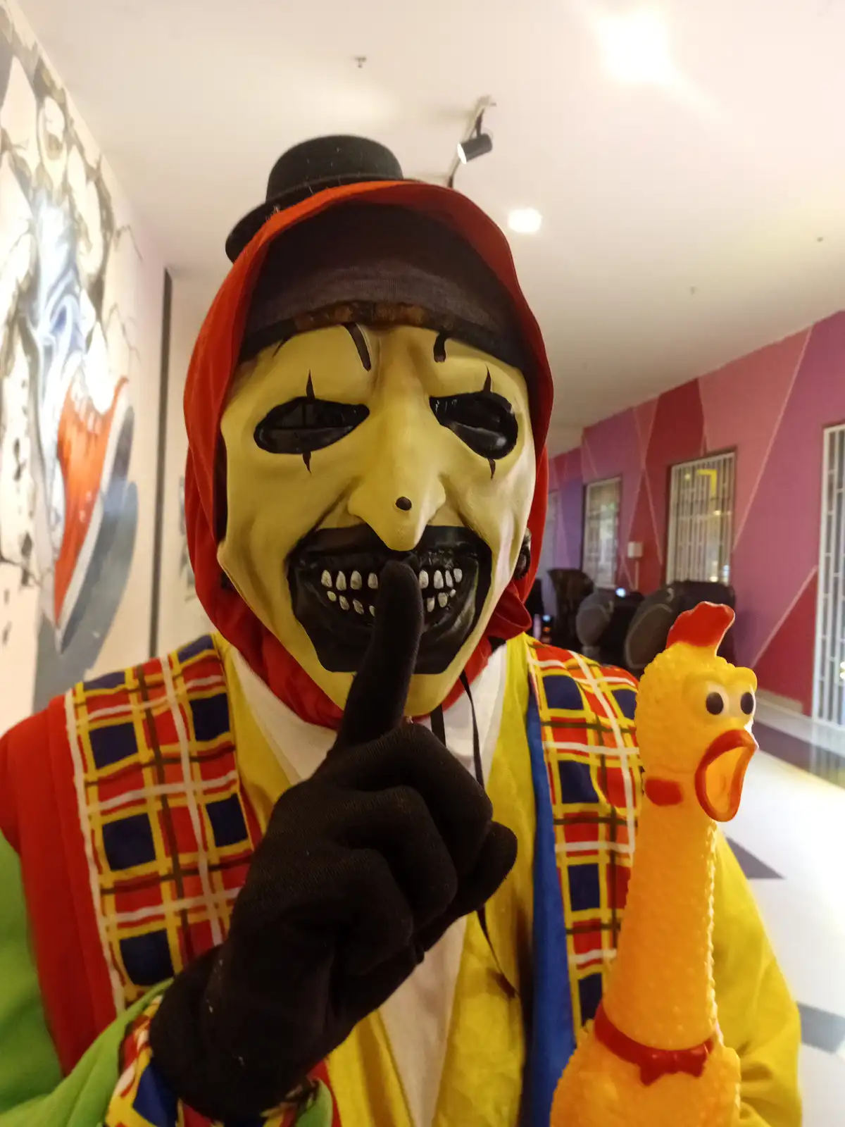 A person wearing a scary yellow clown mask with a top hat and red hood, dressed in a colorful plaid outfit, holds up a…