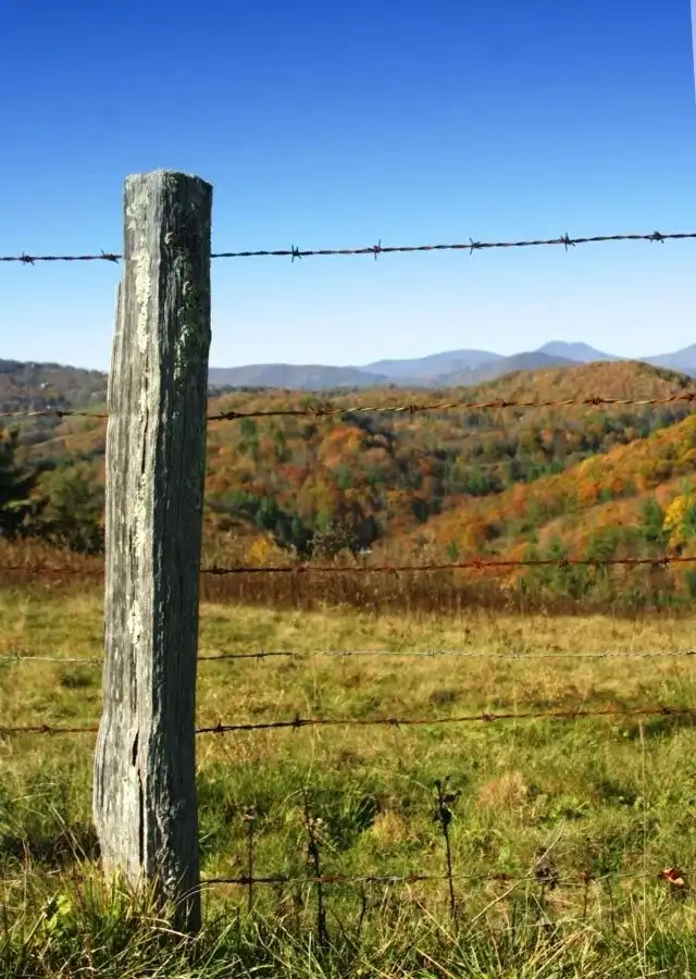 A weathered wooden fence post with barbed wire stretches across a grassy field, overlooking rolling hills covered in autumn…
