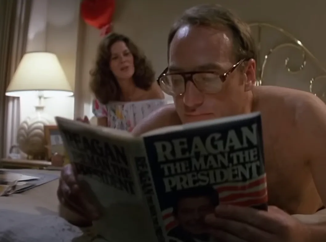 A shirtless man with glasses is lying in bed reading a book titled 'Reagan: The Man, The President' while a woman stands…