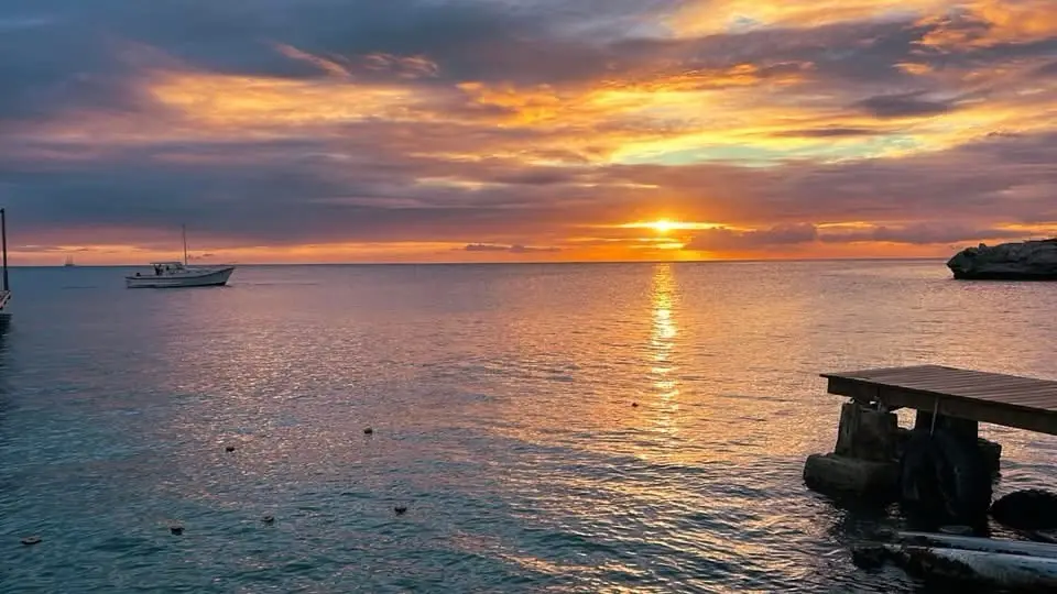 A serene sunset over calm ocean water with a small boat floating in the distance and a wooden pier on the right. The sky is…