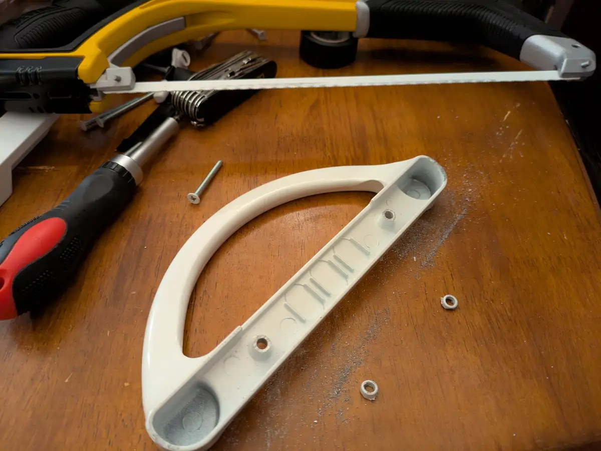 A white plastic door handle component lies on a wooden workbench surrounded by tools including a yellow hacksaw…